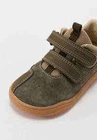 Zapato de ante verde oscuro con parte superior de malla marrón, dos correas de velcro y suela de goma marrón con perforaciones para transpirabilidad.