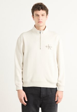 Felpa beige chiaro con collo a zip, polsini a costine e un piccolo logo sul lato sinistro del petto. Realizzata in tessuto morbido e testurizzato.