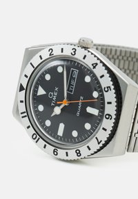 Timex Q ur med et kabinet af rustfrit stål, sort urskive, hvide timemarkeringer, lysende visere og en orange sekundviser, der har et metalmesh-urrem.