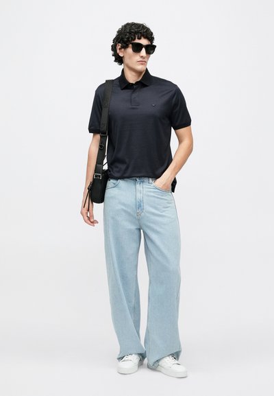 Tumši zila polo krekls ar apkakli, gaiši zili platu kāju džinsi, baltas sporta kurpes un melnas saulesbrilles. Modele nēsā melnu ķermeņa somiņu.