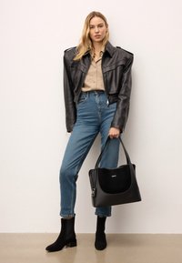 Svart läderhandväska med en slät yta, rundad form och överhandtag. Modellen bär en åtsittande läderjacka, beige skjorta och blå jeans.