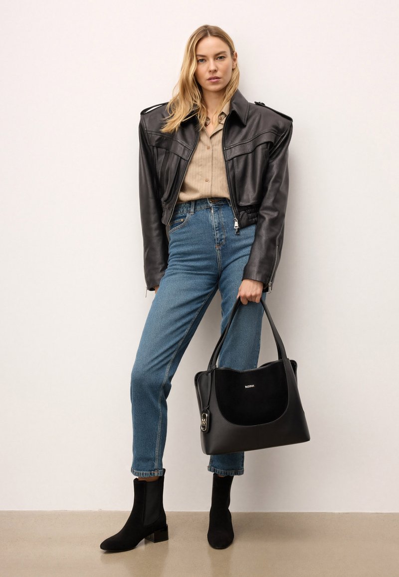 Svart läderhandväska med en slät yta, rundad form och överhandtag. Modellen bär en åtsittande läderjacka, beige skjorta och blå jeans.