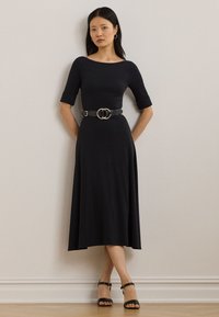 Fekete ruha háromnegyedes ujjakkal, texturált anyagból, derékban megkötve egy dupla gyűrűs csattal ellátott fekete övvel, fekete magassarkú szandálokkal párosítva.