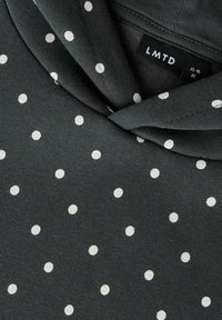 Mørkegrå stoff med hvite polkadotter nærbilde, med en vridd knutedetalj og en synlig svart etikett med teksten "LMTD" og størrelse "15-16 M".