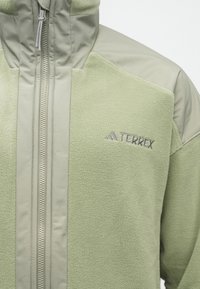 Jachetă din polar verde deschis cu fermoar pe toată fața, material întărit pe umeri și glugă, și logo-ul TERREX pe piept.