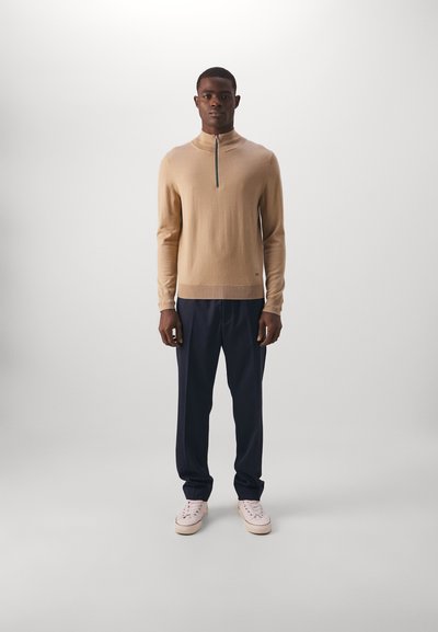 Beige zip-sweater med høj krave, olivenfarvet lynlås og slim fit. Sammen med navy bukser og lyserøde sneakers.