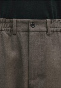 Pantalon en laine gris avec taille élastique, passants pour ceinture et fermeture par bouton noir, porté avec un top noir.