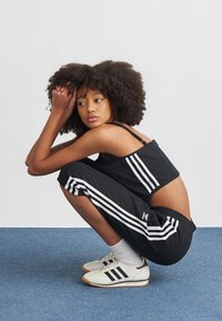 Zwart atletisch ensemble met witte strepen, bestaande uit een cropped top en taps toelopende broek. Model draagt witte sneakers met zwarte strepen, op blauwe achtergrond.