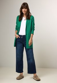 Zelený blazer přes bílý top, doplněný širokými tmavě modrými džíny a béžovými mokasíny. Hladká tkanina, uvolněný střih, praktický design.