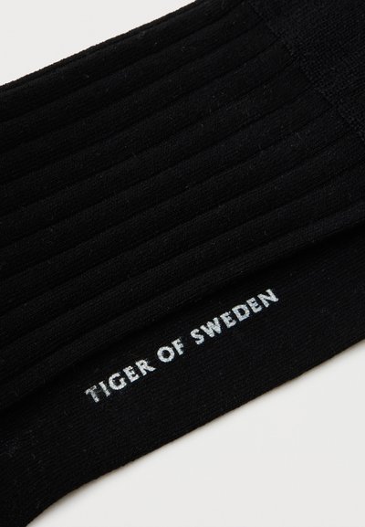Μαύρες ριμπ κάλτσες με την κεντημένη φράση "TIGER OF SWEDEN" σε λευκό χρώμα κατά μήκος της μανσέτας σε λευκό φόντο.