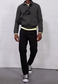 Grau zip-up Bomberjacke, neon-gelber Kragen, schwarze Hose und schwarze Sneakers mit weißen Schnürsenkeln. Kleine Tasche über die Schulter gehängt.