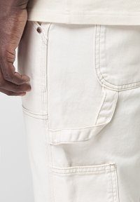 Pantalons cargo beige dotés de plusieurs poches, d'un tissu texturé et d'accents de couture visibles. Poche latérale avec un rabat.