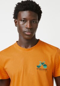 Korte mouwen oranje katoenen t-shirt met een blauwe fietsafbeelding en de tekst "ALTIJD FIETSEN" in het zwart. Glad materiaal, standaard pasvorm.