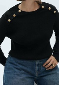 Pull en maille noire avec détails côtelés aux épaules et boutons dorés. Assorti à un jean en denim bleu, présentant une silhouette près du corps.