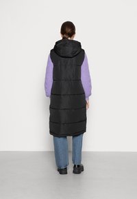 Gilet sans manches noir en duvet avec capuche, doté de coutures horizontales, porté sur un pull en tricot lilas, associé à un jean bleu et des chaussures noires.