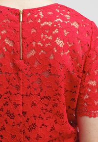 Top in pizzo rosso con motivi floreali, scollo rotondo e cerniera visibile sul retro. La texture appare delicata e intricatata.