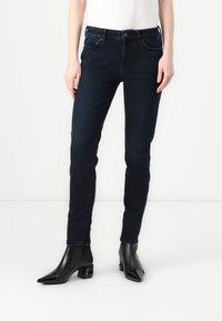 Jeans ajustados de color azul oscuro hechos de denim, con una cintura de altura media, bolsillos delanteros y traseros, y detalles de costura mínimos. Combinados con botas negras de tacón.