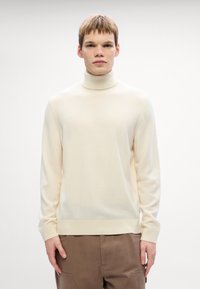 Pulover crema cu guler înalt dintr-un material moale, cu mâneci lungi și manșete striate. Asortat cu pantaloni maro, modelul stă pe un fundal simplu.