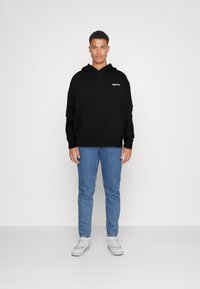 Svart hettegenser med frontlomme og hvit tekstlogo. Kombinert med lyseblå jeans og hvite sneakers. En enkel hvit bakgrunn.