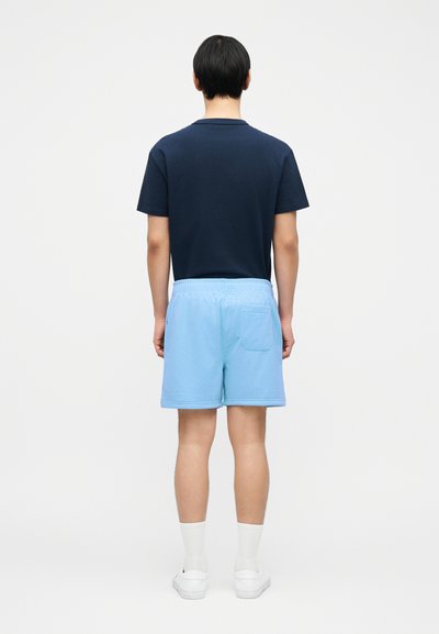 Άνδρας που φοράει μπλε μπλουζάκι και ανοιχτό μπλε σορτς από δίχτυ, λευκές κάλτσες και λευκά αθλητικά παπούτσια, στέκεται με την πλάτη προς τα εμάς σε απλό φόντο.