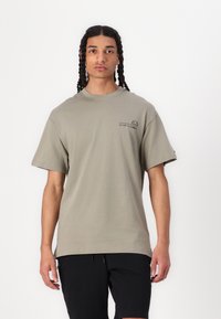 Camiseta de algodón verde oliva, ajuste oversized, cuello redondo, mangas cortas. En la parte frontal presenta un texto negro y un diseño gráfico, estilo minimalista.