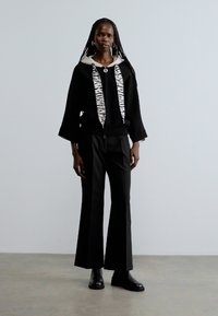 Suéter negro oversize con capucha blanca y negra con patrón, combinado con pantalones negros de pierna ancha y botines negros. Diseño minimalista.