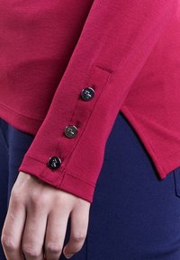 Röd långärmad tröja med rundad nederkant och fyra silverknappar vid manschetten. Slät bomullstyg med avslappnad passform.