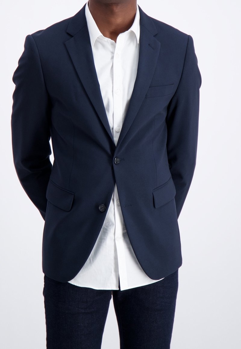 Uomo che indossa un blazer blu scuro su una camicia bianca con pantaloni scuri, in piedi con le mani dietro la schiena su uno sfondo bianco.