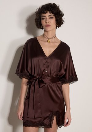 Femme aux cheveux courts et bouclés portant une robe de chambre en satin marron foncé avec bordure en dentelle et ceinture à la taille, debout devant un fond clair uni.