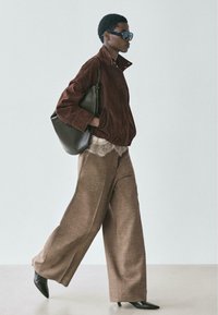 Bruine suède jas, top met kantdetails, brede bruine broek en zwarte puntige laarzen. Donkere zonnebril en een grote zwarte handtas maken de look af.