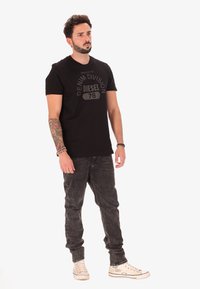 T-shirt in cotone nero con maniche corte, caratterizzato da una stampa grafica in rilievo che recita "PROPRIETÀ DELLA DIVISIONE DENIM DIESEL 78" in grigio. Indossato con jeans scuri.