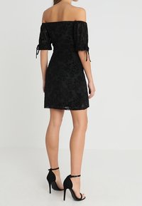 Vestido negro de hombros descubiertos con mangas cortas abullonadas, patrón de encaje floral, cintura ajustada y falda acampanada. Combinado con sandalias negras de tacón alto.
