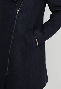 Marinblå kappa i fiskbensmönster med silvrig dragkedja och sidan ficka, hand delvis i fickan, mot en enfärgad ljus bakgrund.