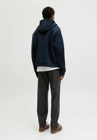 Marinblå hoodie med stor huva, avslappnad passform och ribbade muddar, kombinerad med grå byxor och bruna skor, sedd bakifrån.