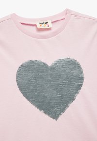 Vaaleanpunainen puuvillapaita, jossa on suuri hopeanvärisistä paljeteista valmistettu sydäntulo etupuolella, pyöreä kaula-aukko ja lyhyet hihat.