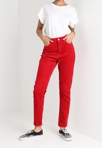 Pantaloni in corduroy rossi con vita alta, dotati di chiusura frontale con bottone e vestibilità slim, abbinati a una maglietta bianca e sneakers nere.