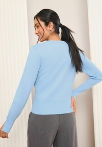 Ljusblå, långärmad stickad tröja med ribbad midja och manschetter, bärs tillsammans med grå, löst sittande byxor.