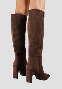 Bottes en suédine marron jusqu'au genou avec un talon haut épais, un bout pointu et un détail de fermeture éclair sur le côté. Texture lisse avec une silhouette élégante.