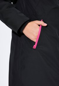 Musta ulkovaatteet, joissa on vetoketjullinen tasku kirkkaan pinkillä vetoketjulla. Kangas on sileäpintaista, sopivaa erilaisiin sääolosuhteisiin.