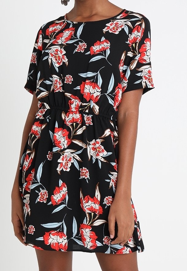 Femme portant une robe noire à manches courtes avec un motif floral rouge, blanc et bleu et une taille élastique, debout devant un fond blanc uni.