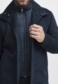 Tamna vunena kaput s dugmadi sprijeda, s podstavom od sjajne tkanine i visokim ovratnikom. Vidljiva tekstura i detalj sa patentnim zatvaračem.