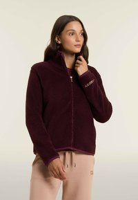 Donna che indossa una giacca in pile bordeaux con cerniera, abbinata a pantaloni beige chiaro con cordoncino, che guarda leggermente a destra su uno sfondo neutro.