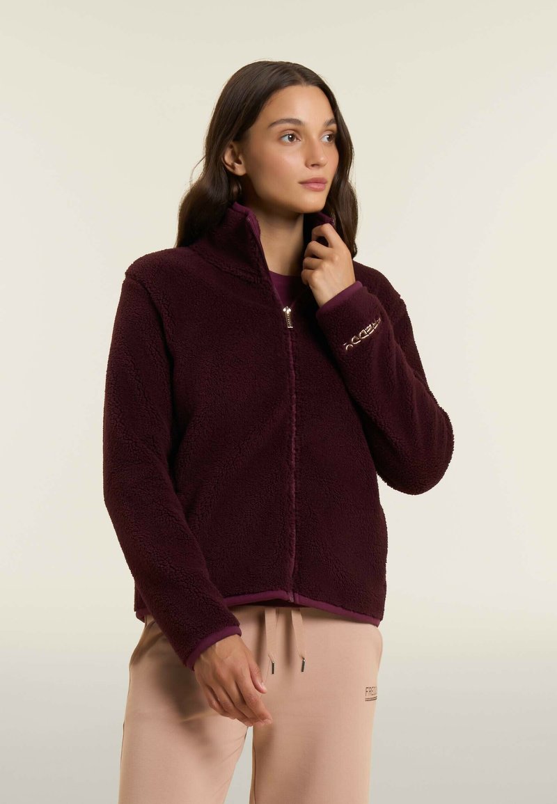 Donna che indossa una giacca in pile bordeaux con cerniera, abbinata a pantaloni beige chiaro con cordoncino, che guarda leggermente a destra su uno sfondo neutro.