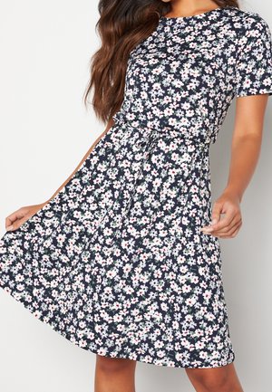Woman wearing a short-sleeve, navy blue dress with white and pink floral print and a tied waist belt, holding the dress skirt with one hand.