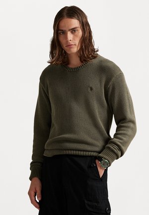 Giovane uomo con capelli castani che arrivano alle spalle, che indossa un maglione a maglia verde oliva e pantaloni cargo neri, con una mano in tasca.