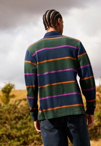 Pruhovaný pulovr v tmavě zelené barvě, s horizontálními pruhy v oranžové, fialové a modré. Dlouhé rukávy, pletená textura, límcový střih.