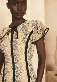 Kleid aus einem transparenten, gemusterten Stoff in Gelb-, Schwarz- und Brauntönen, mit Rüschenschlaufen und einem Bindeband am Hals.