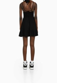 Černé bezramínkové šaty se smockovaným zadním dílem a volánkovým lemem, doplněné černo-bílými teniskami ve vysokém provedení.