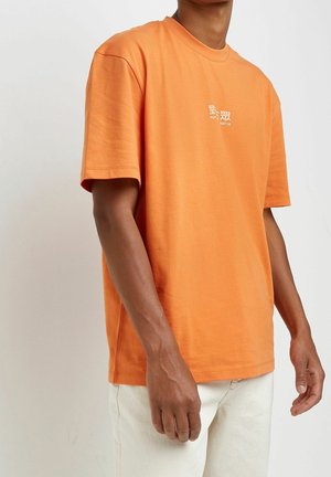 Orangenes T-Shirt mit kurzen Ärmeln aus Baumwolle, mit einem Rundhalsausschnitt und minimalistischem weißen Textdesign auf der Vorderseite. Kombiniert mit hellen Hosen.
