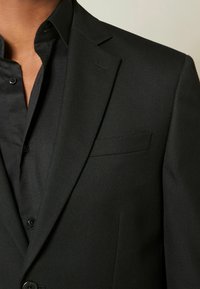 Blazer noir texturé avec un revers cranté, une poche poitrine simple, et une chemise noire boutonnée visible en dessous.
